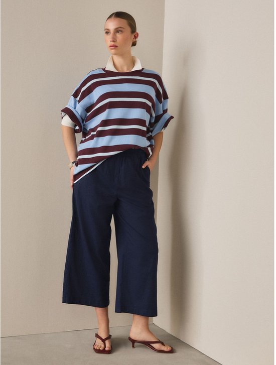 Model draagt donkerblauwe wijde broek met elastische tailleband, gecombineerd met gestreept shirt en bruine sandalen.