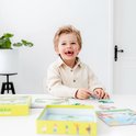 Een lachende jongen met blond haar zit aan tafel en puzzelt. Op tafel liggen puzzels met een kikker en andere dieren.