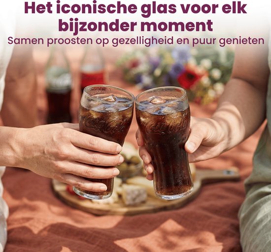 Deux mains lèvent un verre Coca-Cola avec du cola et des glaçons pour trinquer, avec une couverture de pique-nique et de la nourriture en arrière-plan.