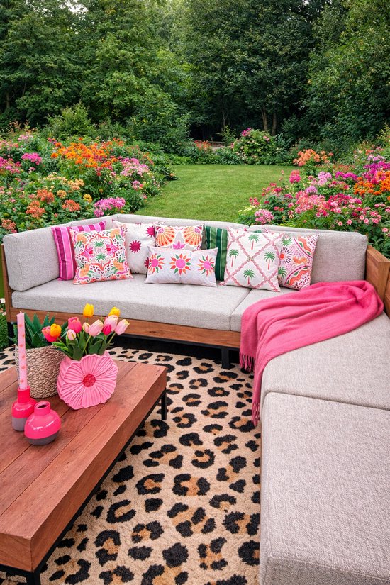 Lichtbruine hoekbank in een tuin, vol met kleurrijke sierkussens, een roze plaid en een luipaardprint vloerkleed.
