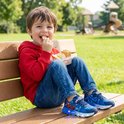 Jongen in rood vest en spijkerbroek zit op een bankje in het park, met een lunchtrommel. Hij draagt blauwe Spiderman schoenen met lichtjes.