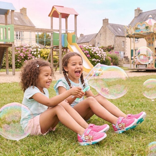 Twee lachende meisjes met roze sneakers met Paw Patrol print, zittend op gras met bellenblaas.