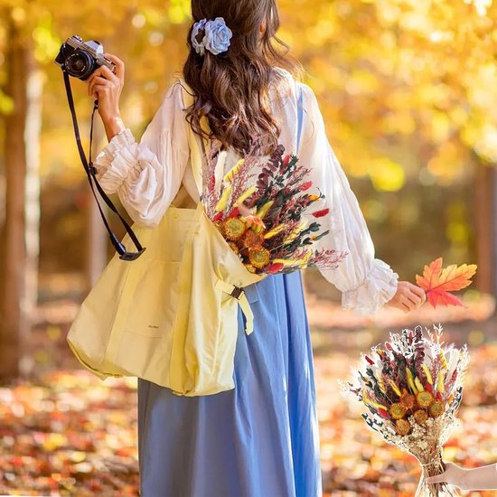 Vrouw in een blauwe jurk en witte blouse houdt een tas met een herfstboeket vast in een bos.
