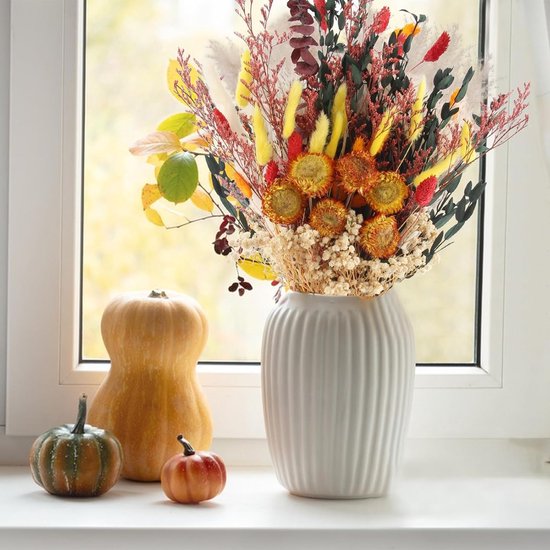 Herfstboeket van droogbloemen in een witte geribbelde vaas op een vensterbank met kleine pompoenen.