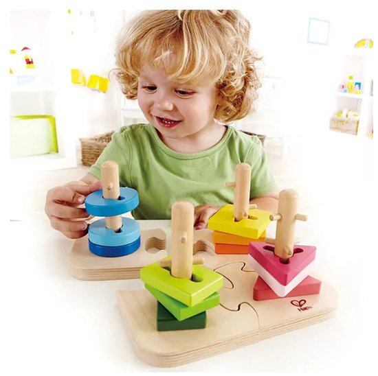 Kind met krullend haar speelt met de Hape vormpuzzel. Stapelt blauwe, groene, roze en gele vormen op houten pegs.