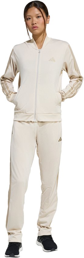 Vrouw met trainingspak: crèmekleurig jack met beige strepen en beige trainingsbroek, beide met Adidas logo.