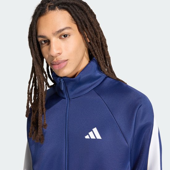 Close-up van een man met dreadlocks die een blauw trainingsjack draagt met witte accenten.