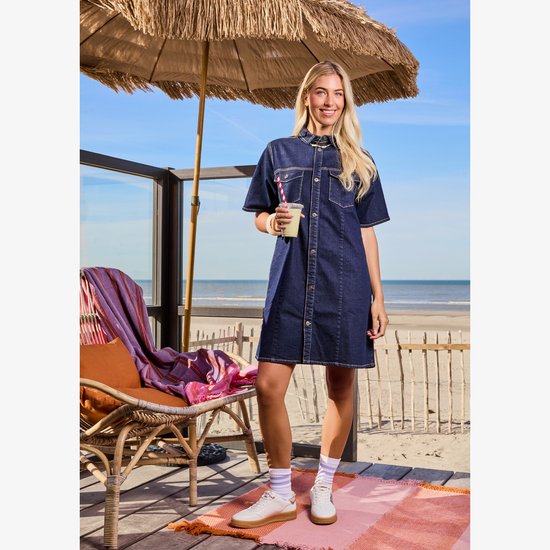 Vrouw met blond haar in een donkerblauwe spijkerjurk met korte mouwen, op het strand.