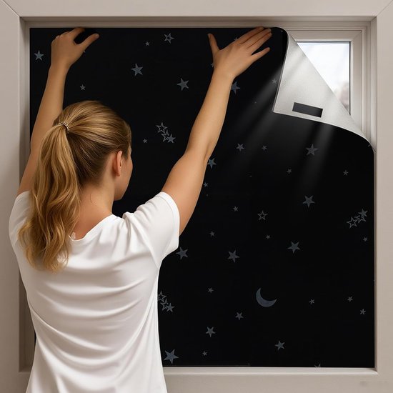 Une femme applique un rideau occultant noir avec un motif d'étoiles et de lunes sur une fenêtre.