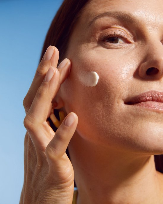 Close-up van een vrouw die witte crème aanbrengt op haar wang onder een blauwe hemel.