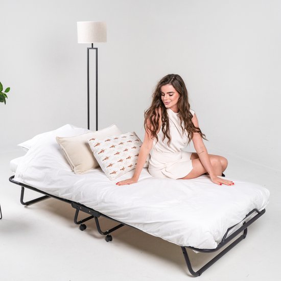 Wit opklapbed met een opgerold matras, twee kussens en een vrouw die erop zit. Een staande lamp staat naast het bed.