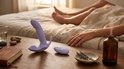 Paarse vibrator en afstandsbediening op een tafel naast een bed waar koppels liggen, met kaars en glas water.