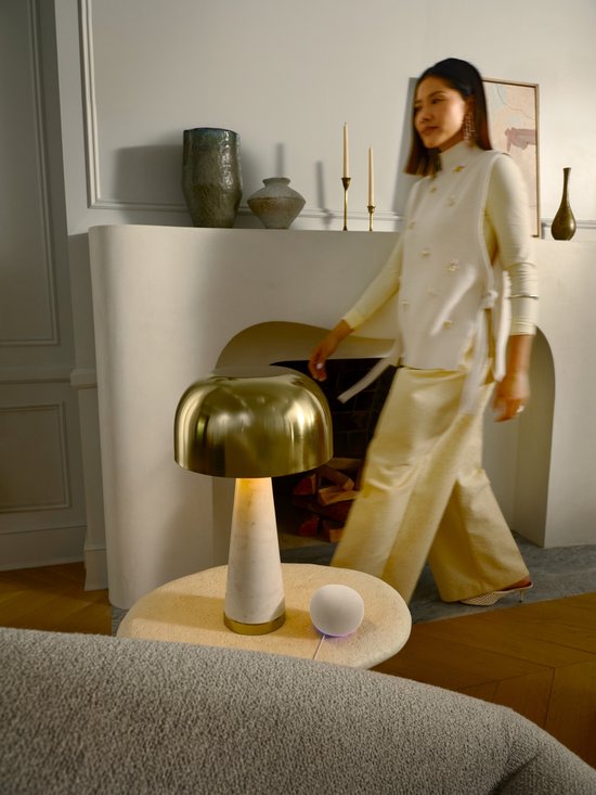 Witte Google Home speaker met gekleurde gloed, naast een bijzettafel met lamp.