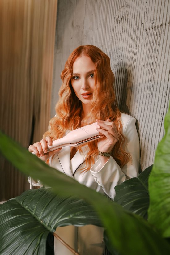 Vrouw met lang, golvend rood haar houdt een roze haarkruller vast, gekleed in een wit jasje.