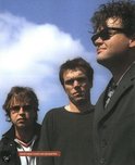 Drie mannen, waarvan de rechter met zonnebril, kijken omhoog tegen een blauwe lucht.
