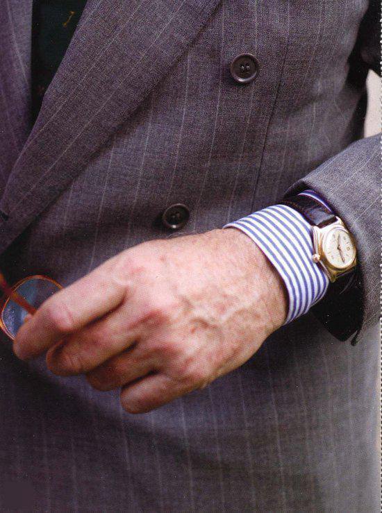 Man in grijs gestreept pak, met blauw-wit gestreepte manchet en gouden horloge.