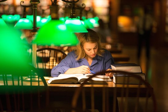 Een blonde vrouw in een blauw shirt leest geconcentreerd in een dik boek in een bibliotheek.