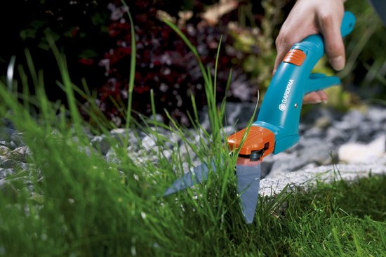 Hand maait gras naast planten met blauwe Gardena graschaar.