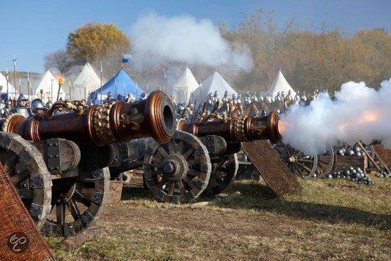 Twee kanonnen op wielen vuren af met rook, met een tentenkamp en soldaten op de achtergrond.
