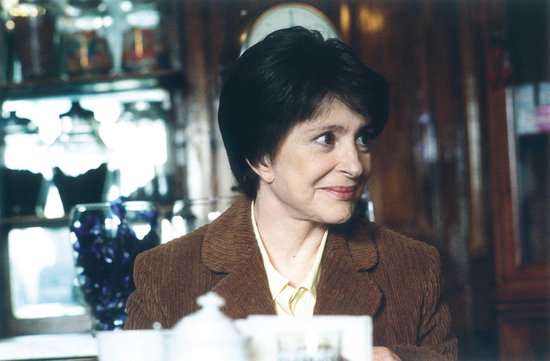 Een vrouw met zwart haar en een bruine corduroy jas kijkt lachend weg. Ze draagt een gele blouse.