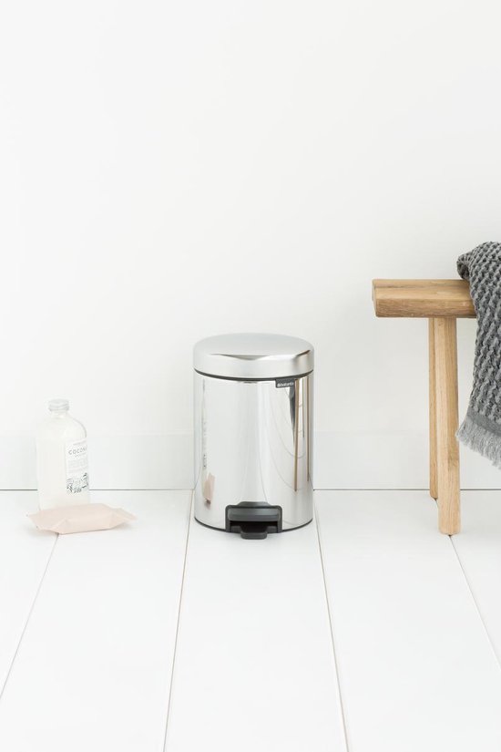 Briljant stalen Brabantia NewIcon prullenbak met zwart voetpedaal, naast een houten bankje met handdoek.