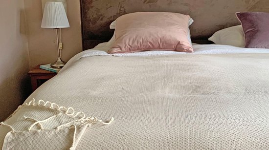 Een zandkleurig plaid met franjes ligt op een bed met een roze kussen.
