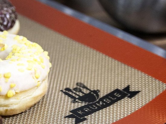 Donut met witte glazuur en gele spikkels op een beige bakmat met bruine rand en Krumble logo.