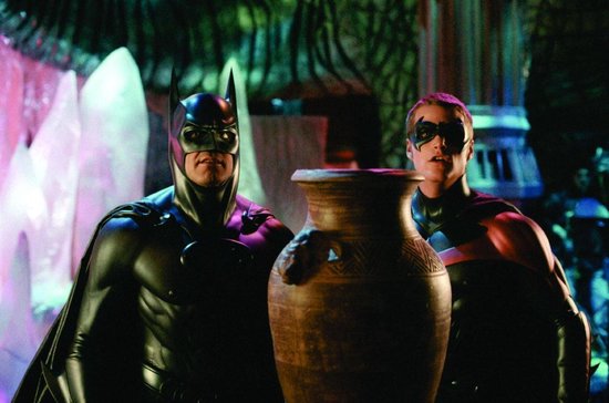 Batman en Robin staan naast een grote vaas in een verlichte, donkere ruimte. Ze dragen hun iconische kostuums.