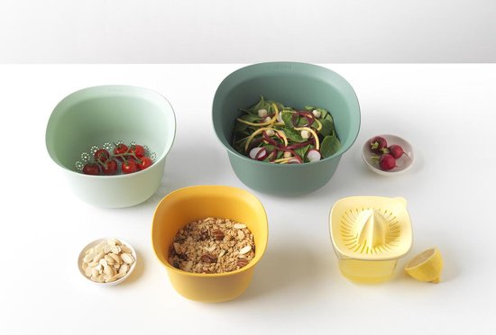 Een gele beslagkom met muesli, een groene kom met salade, een lichtgroene geperforeerde kom met kerstomaatjes en een gele citruspers.