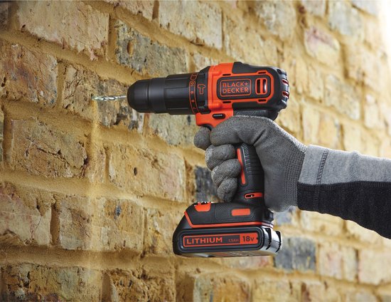 Hand met grijze handschoen die een oranje en zwarte BLACK+DECKER boormachine gebruikt om in een bakstenen muur te boren.