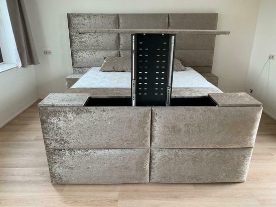 Grijze boxspring met TV-lift, met gestoffeerd hoofdbord en bijzettafels.