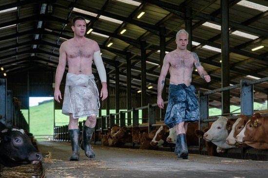 Twee mannen, half ontbloot en met doeken omgeslagen, lopen in een stal tussen koeien.