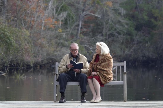 Een ouder stel zit op een bankje aan het water. De man leest een boek, de vrouw luistert. De man draagt een beige jas en de vrouw een oranje sjaal.
