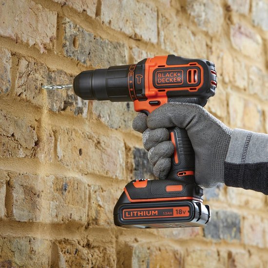 Een hand met een grijze werkhandschoen houdt een zwarte en oranje Black+Decker klopboormachine vast. De boormachine boort in een bakstenen muur.