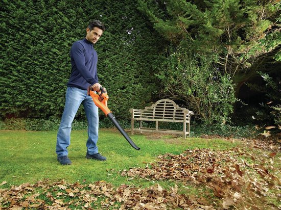 Man in blauwe trui en spijkerbroek gebruikt oranje bladblazer in tuin met herfstbladeren.