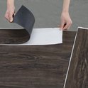 Handen die de beschermlaag van een donkerbruine PVC vloerplank met houtnerf verwijderen.