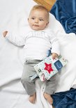 Baby met blauwe ogen ligt op een witte deken en houdt een stoffen boekje vast.