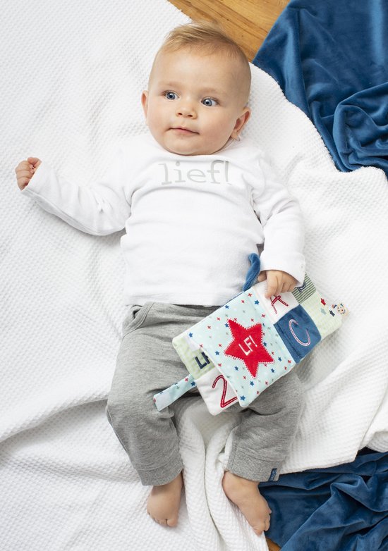 Baby met blauwe ogen ligt op een witte deken en houdt een stoffen boekje vast.