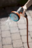 Grijze GARDENA spuit met oranje en blauwe accenten sproeit water op een betegeld pad.