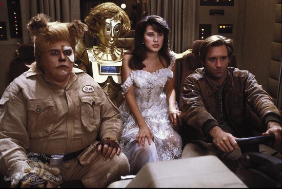 Een grote, harige alien met oren, een gouden robot, een vrouw in een witte jurk en een man in een bruin jack zitten in een cockpit.