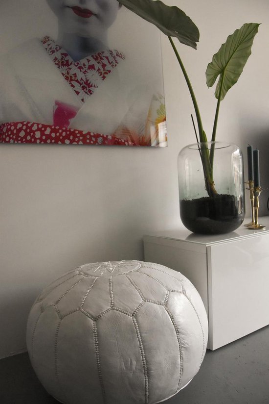 Ronde witte poef met strakke naden en een zilverkleurig detail in het midden, naast een witte kast met planten en kunst.