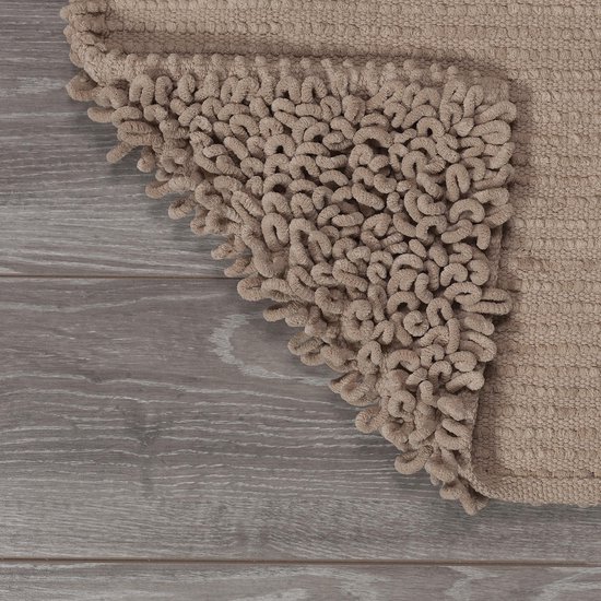 Tapis de bain couleur sable avec des boucles torsadées, partiellement enroulé sur un sol en bois.