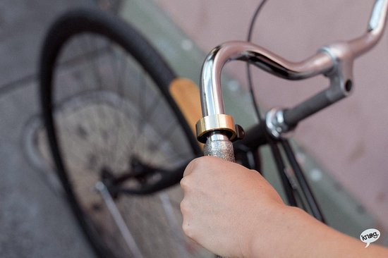 Hand die een zilverkleurige fietsbel met een gouden rand op het stuur van een fiets vastpakt.