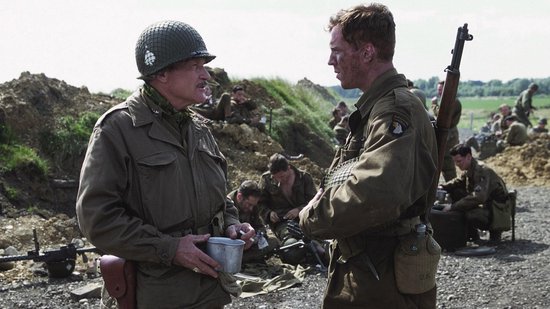 Twee militairen in uniform buiten met een wapen, pratend, met andere soldaten op de achtergrond.