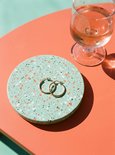 Goudkleurige hoepeloorbellen liggen op een rond, mintgroen terrazzo onderzettertje naast een glas met drankje.