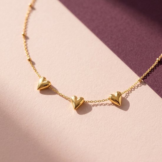 Goudkleurige ketting met drie kleine hartjeshangers op een roze en paarse achtergrond.