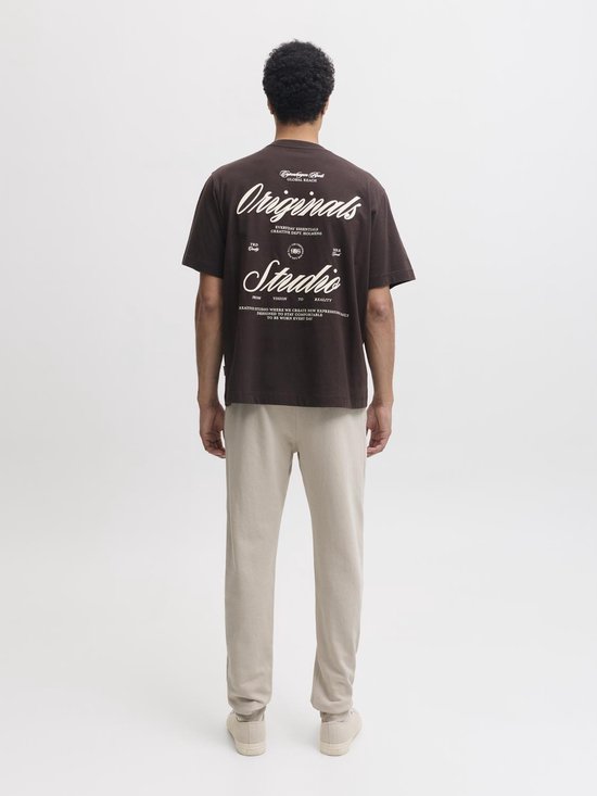 Man in bruin T-shirt met grote witte 'Originals Studio' tekst op de rug.