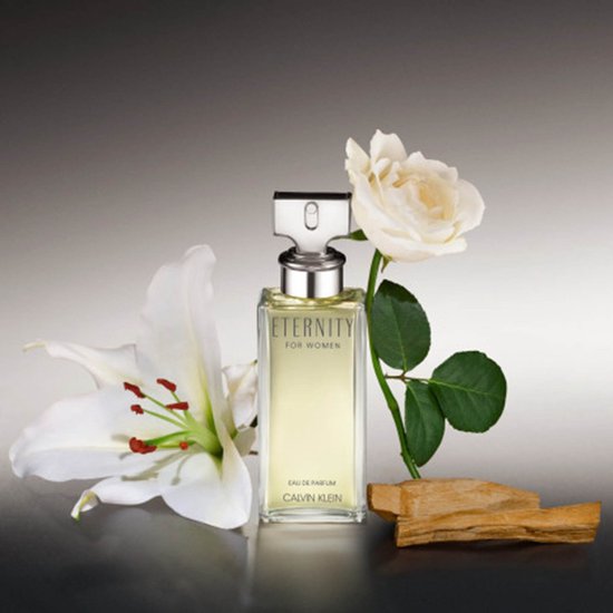 Eternity for Women Eau de Parfum van Calvin Klein, in een heldere glazen flacon met zilveren dop, naast witte lelies en een roos.