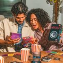 Twee personen spelen Hitster bordspel met 'Movies & TV Soundtracks' uitbreidingsset, popcorn en speelkaarten op tafel.
