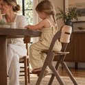 Kind in geel-witte ruitjes jumpsuit zittend op een donkerbruine Deryan houten kinderstoel aan de eettafel.